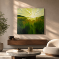 A square canvas art print depicting a vibrant green and yellow landscape with a bright sunburst, displayed on a beige wall in a modern living room setting with a wooden console, armchair, and potted plant.