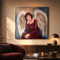 A square canvas painting of a seated angel with large white wings and a deep red robe, displayed on a beige wall in a styled living room with a burgundy armchair, table, and lamp.