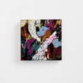 An abstract square canvas painting with textured, impasto brushstrokes in a multicolor palette of purple, blue, white, black, and red, displayed against a clean white wall.